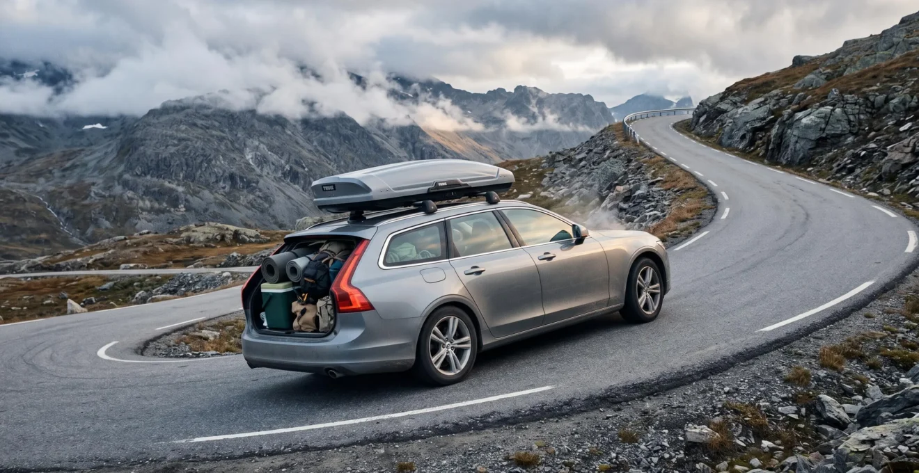 A heavily loaded family car with a roof box climbing a steep paved mountain pass road under partly cloudy skies.